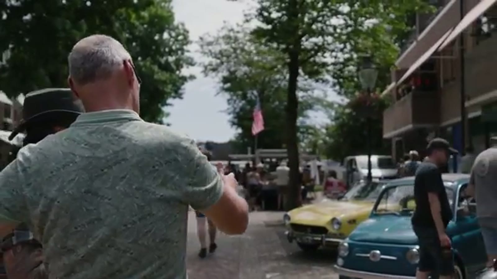 Turquoise Fiat 500 tussen de klassiekers op het dorpsplein van Losser, met publiek en een Amerikaanse vlag op de achtergrond