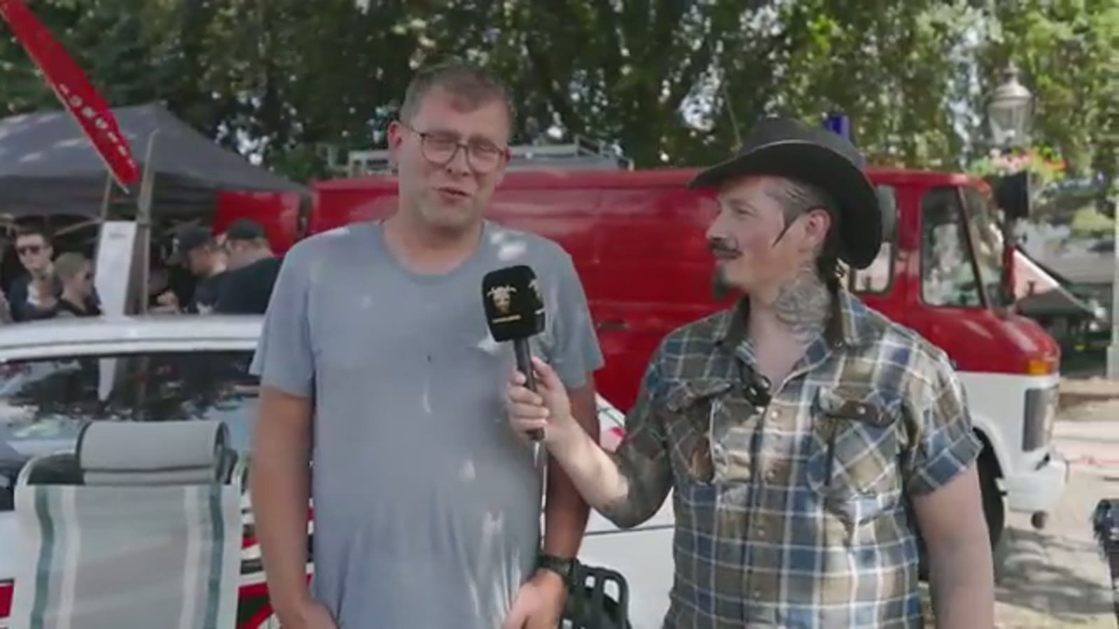 Presentator met cowboyhoed en snor interviewt een standhouder met microfoon op het evenemententerrein in Losser